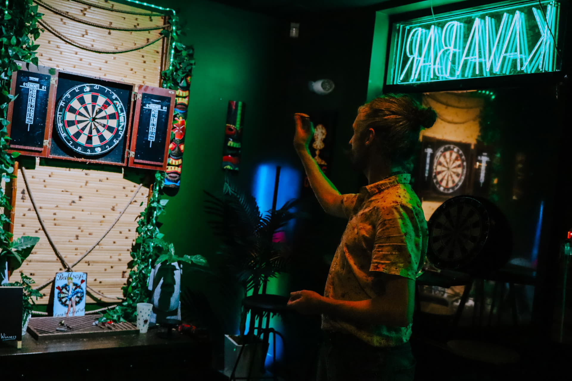 Player throwing at the tiki dartboard under the neon kava bar sign during Thursday darts